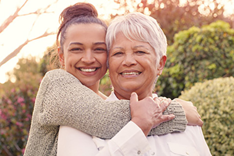 Smiling woman hugging her elderly mother outdoors on a sunny day.