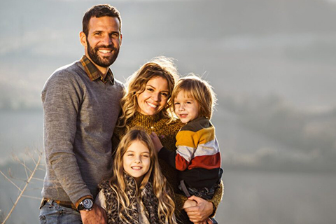 Young family of four smiling outdoors with mountain scenery in the background.